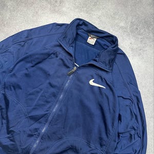 Pode incluir: Casaco de treino azul marinho com z&iacute;per completo e logotipo branco da Nike no peito. O casaco tem gola alta e uma etiqueta perto da gola. O casaco &eacute; feito de um material atl&eacute;tico liso.