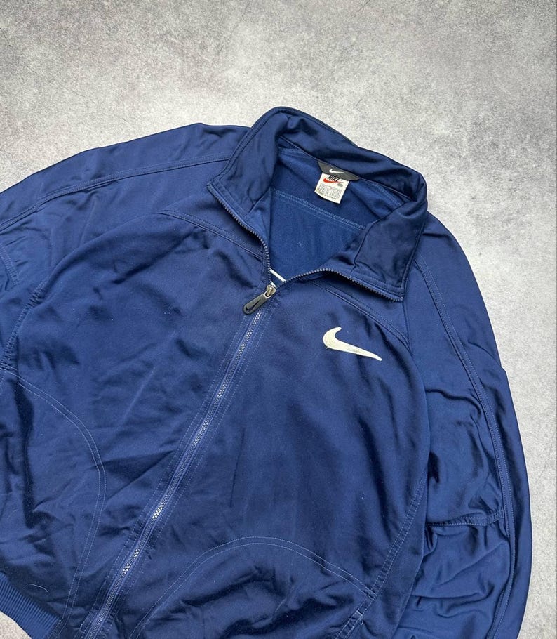 Pode incluir: Casaco de treino azul marinho com z&iacute;per completo e logotipo branco da Nike no peito. O casaco tem gola alta e uma etiqueta perto da gola. O casaco &eacute; feito de um material atl&eacute;tico liso.