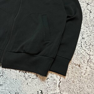 May include: Black zip-up jacket with a small heart-shaped emblem featuring two white eyes. The jacket has a ribbed hem and cuffs, and a single pocket. The fabric appears to be a soft, medium-weight material.