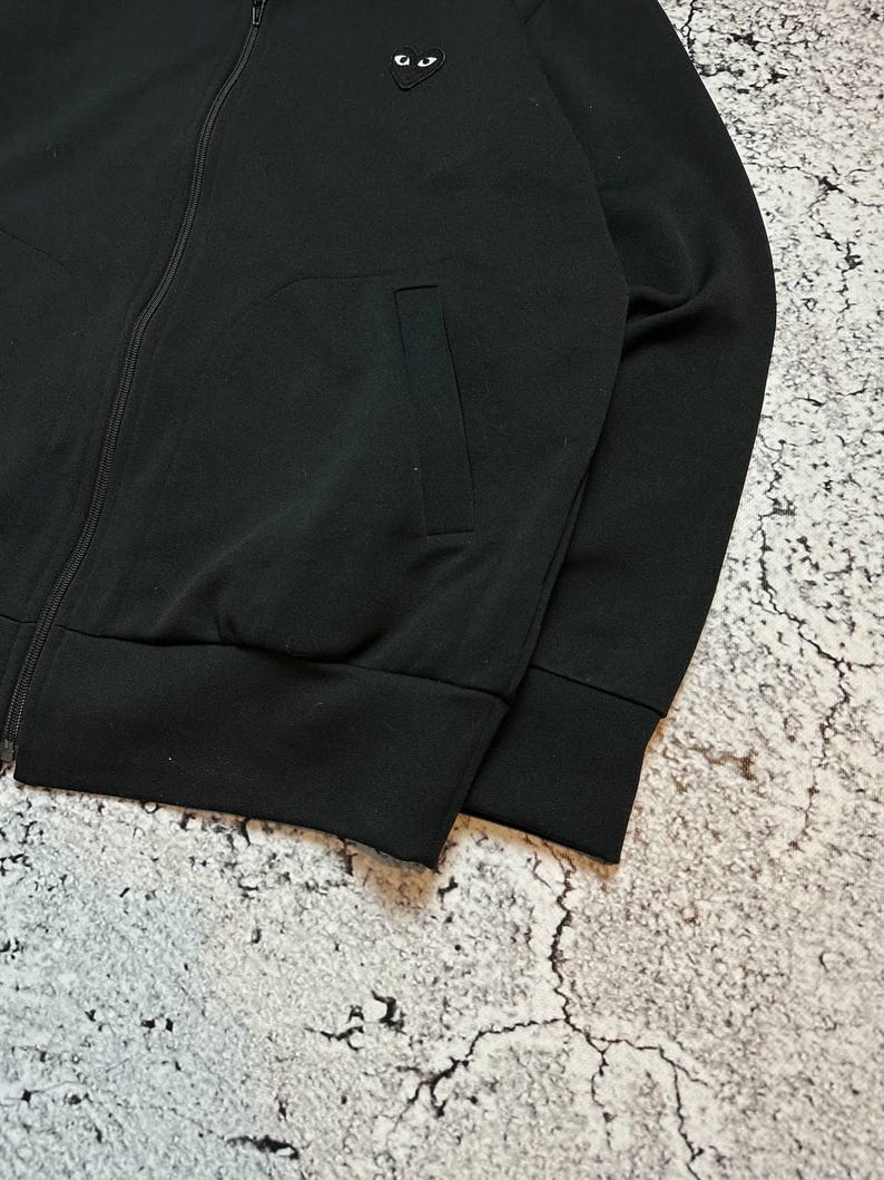 May include: Black zip-up jacket with a small heart-shaped emblem featuring two white eyes. The jacket has a ribbed hem and cuffs, and a single pocket. The fabric appears to be a soft, medium-weight material.