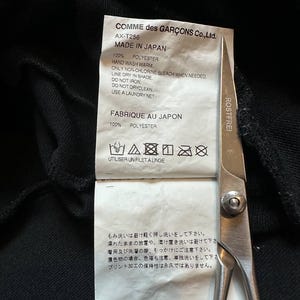 May include: A close-up of a black garment tag with text in English and Japanese. The tag reads "COMME des GAR&Ccedil;ONS Co., Ltd." and "MADE IN JAPAN." Scissors are in the frame. The tag also includes washing instructions.