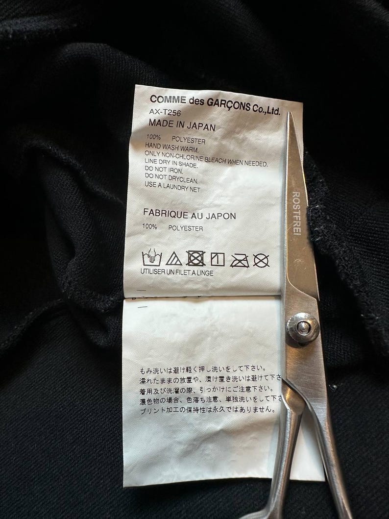 May include: A close-up of a black garment tag with text in English and Japanese. The tag reads "COMME des GAR&Ccedil;ONS Co., Ltd." and "MADE IN JAPAN." Scissors are in the frame. The tag also includes washing instructions.