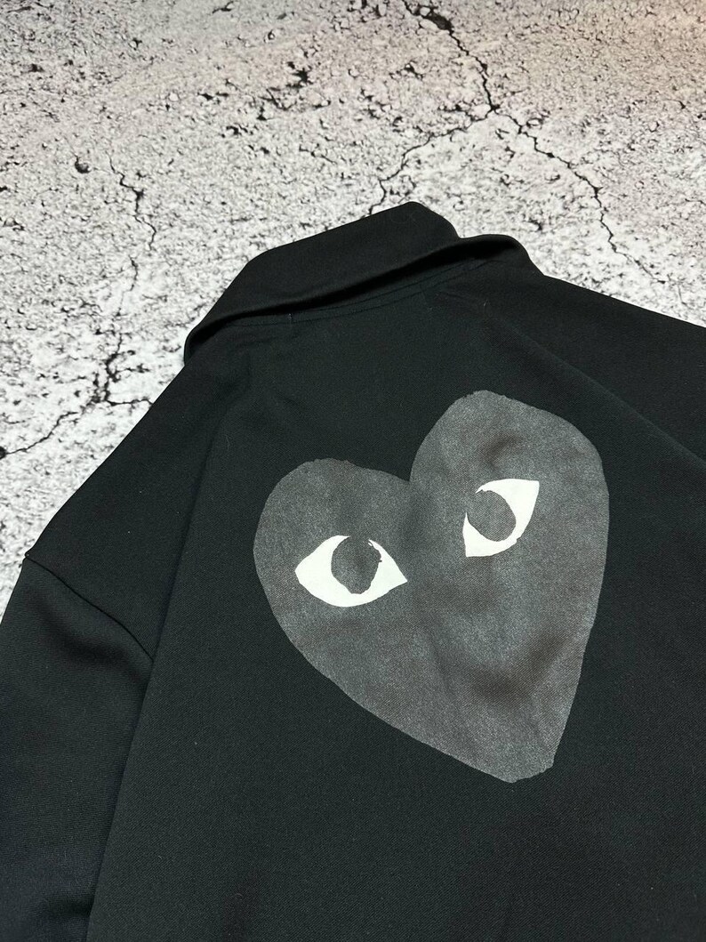 May include: Black sweatshirt featuring a large heart-shaped graphic with white eyes on the back. The sweatshirt has a collar and is made of a soft material. The graphic is centered on the back of the garment.