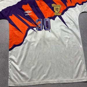 May include: White soccer jersey with purple and orange accents. Features the number 30, the Umbro logo, and a Scottish Football Association emblem. The jersey has a collared neckline.
