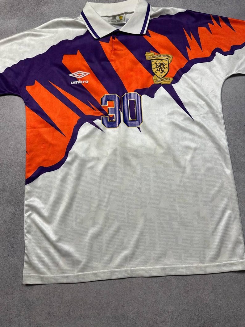 May include: White soccer jersey with purple and orange accents. Features the number 30, the Umbro logo, and a Scottish Football Association emblem. The jersey has a collared neckline.