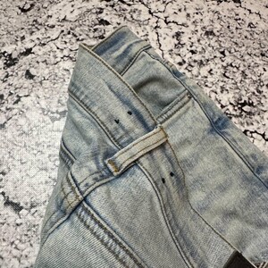 Op de afbeelding: Close-up van een lichtblauwe spijkerbroek. De jeans heeft een klassiek ontwerp met vijf zakken, riemlussen en een knoop- en ritssluiting. De denim heeft een vervaagde, versleten uitstraling met zichtbare stiksels.