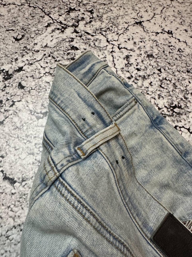 Op de afbeelding: Close-up van een lichtblauwe spijkerbroek. De jeans heeft een klassiek ontwerp met vijf zakken, riemlussen en een knoop- en ritssluiting. De denim heeft een vervaagde, versleten uitstraling met zichtbare stiksels.