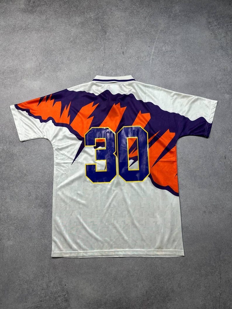 May include: White athletic jersey with a purple and orange design across the shoulders and back. The number "30" is prominently displayed on the back in blue with a gold outline. The jersey has a collared neckline.