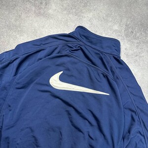 Pode incluir: Casaco desportivo azul marinho com o log&oacute;tipo branco da Nike nas costas. O casaco tem gola alta e fecho de correr. O material parece ser uma mistura leve, possivelmente nylon ou poli&eacute;ster.