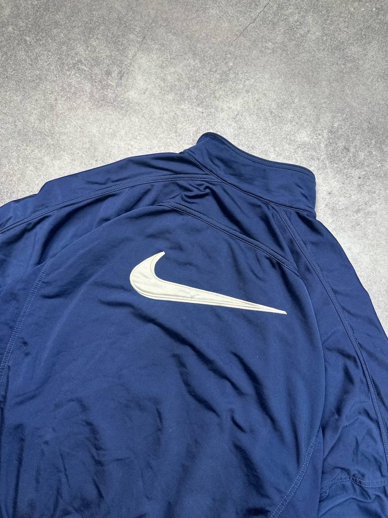 Pode incluir: Casaco desportivo azul marinho com o log&oacute;tipo branco da Nike nas costas. O casaco tem gola alta e fecho de correr. O material parece ser uma mistura leve, possivelmente nylon ou poli&eacute;ster.