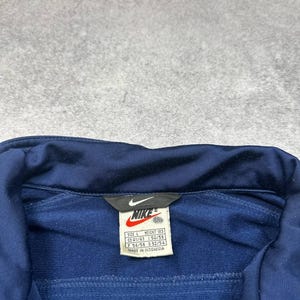 Pode incluir: Casaco de treino Nike azul marinho com uma etiqueta branca com o log&oacute;tipo da Nike e informa&ccedil;&otilde;es de tamanho. O casaco tem um fecho de correr e uma gola. A etiqueta diz "MADE IN INDONESIA".