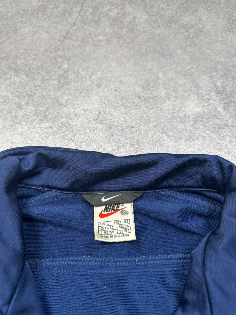 Pode incluir: Casaco de treino Nike azul marinho com uma etiqueta branca com o log&oacute;tipo da Nike e informa&ccedil;&otilde;es de tamanho. O casaco tem um fecho de correr e uma gola. A etiqueta diz "MADE IN INDONESIA".