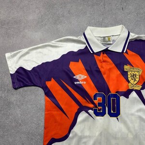 May include: White, purple, and orange soccer jersey with the number 30 in blue. The jersey has a collared neckline and the Umbro logo. The Scottish Football Association emblem is on the right chest.