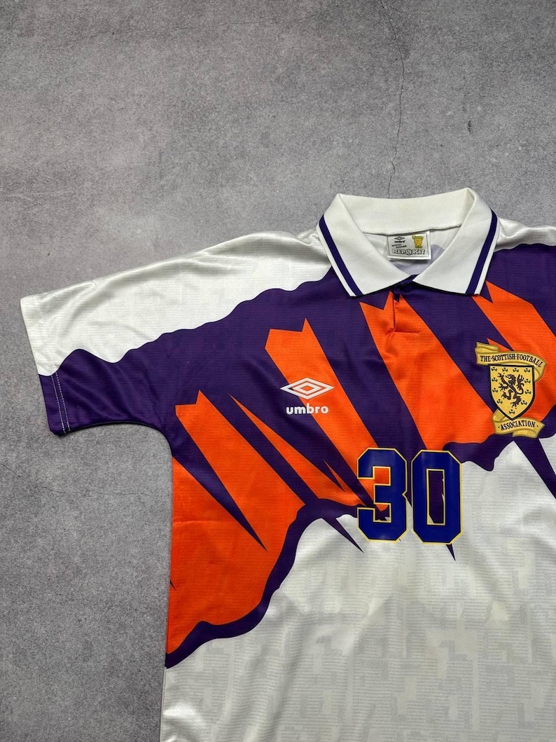 May include: White, purple, and orange soccer jersey with the number 30 in blue. The jersey has a collared neckline and the Umbro logo. The Scottish Football Association emblem is on the right chest.