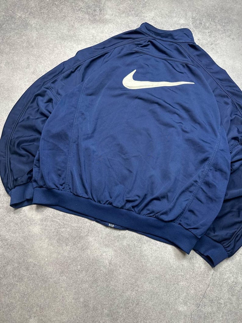 Pode incluir: Casaco desportivo azul marinho com o log&oacute;tipo Nike branco nas costas. O casaco tem fecho de correr completo, punhos canelados e bainha canelada. O casaco &eacute; feito de um material leve e respir&aacute;vel.