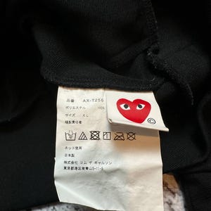May include: Close-up of a black garment tag with Japanese text and a red heart logo with eyes. The tag includes washing instructions and the text "AX-T256" and "XL". The tag is attached to a black fabric.