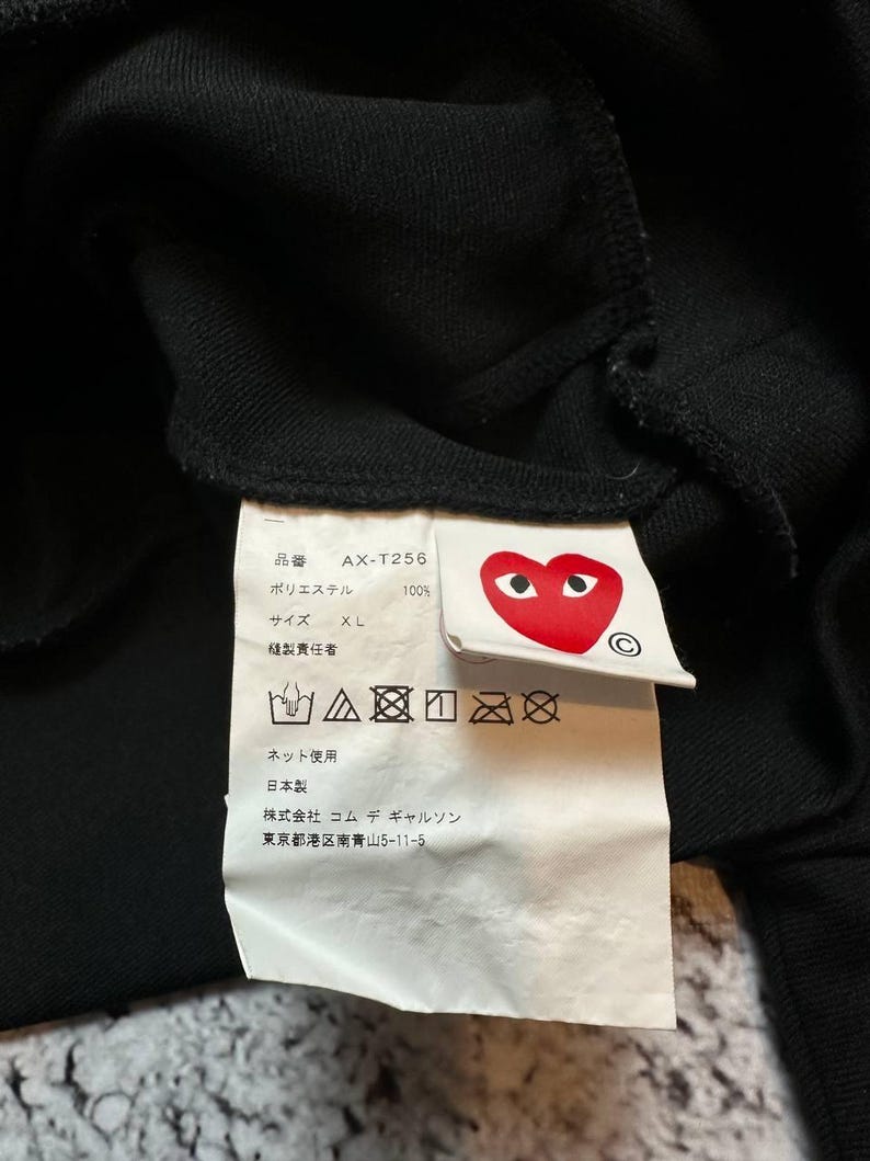 May include: Close-up of a black garment tag with Japanese text and a red heart logo with eyes. The tag includes washing instructions and the text "AX-T256" and "XL". The tag is attached to a black fabric.