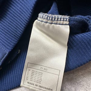 Pode incluir: Close-up de uma pe&ccedil;a de roupa azul marinho com uma etiqueta branca. A etiqueta tem o texto "FT SCI" e "100% POLYESTER" em v&aacute;rias l&iacute;nguas. A pe&ccedil;a de roupa &eacute; feita na Indon&eacute;sia.