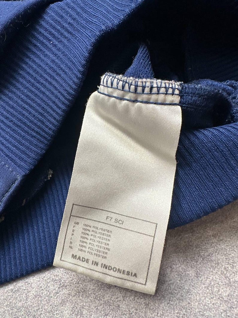 Pode incluir: Close-up de uma pe&ccedil;a de roupa azul marinho com uma etiqueta branca. A etiqueta tem o texto "FT SCI" e "100% POLYESTER" em v&aacute;rias l&iacute;nguas. A pe&ccedil;a de roupa &eacute; feita na Indon&eacute;sia.