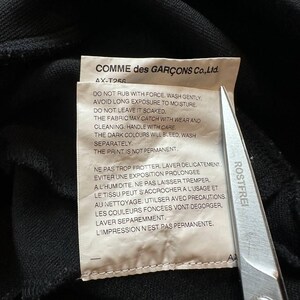 May include: A close-up of a black garment with a care label. The label reads "COMME des GAR&Ccedil;ONS Co., Ltd." and includes washing instructions in English and French. A pair of scissors is partially visible.