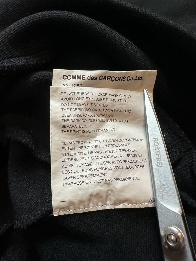 May include: A close-up of a black garment with a care label. The label reads "COMME des GAR&Ccedil;ONS Co., Ltd." and includes washing instructions in English and French. A pair of scissors is partially visible.