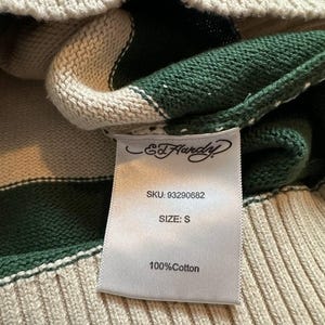 Pu&ograve; includere: Primo piano di un maglione a righe verdi e crema. L'etichetta recita "Ed Hardy" e include lo SKU, la taglia "S" e "100% Cotone". Il maglione ha una trama a coste.