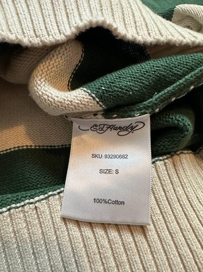 Pu&ograve; includere: Primo piano di un maglione a righe verdi e crema. L'etichetta recita "Ed Hardy" e include lo SKU, la taglia "S" e "100% Cotone". Il maglione ha una trama a coste.