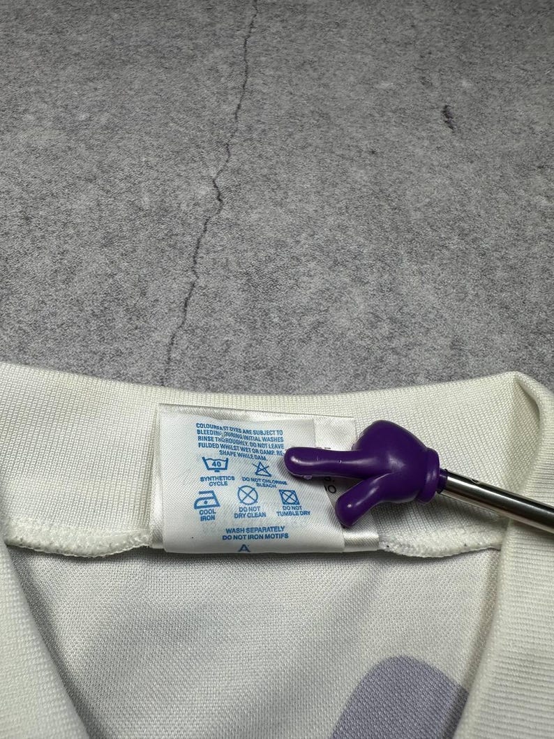 May include: Close-up of a white garment tag with care instructions. The tag includes symbols for washing, drying, and ironing. A purple hand-shaped pointer is touching the tag.