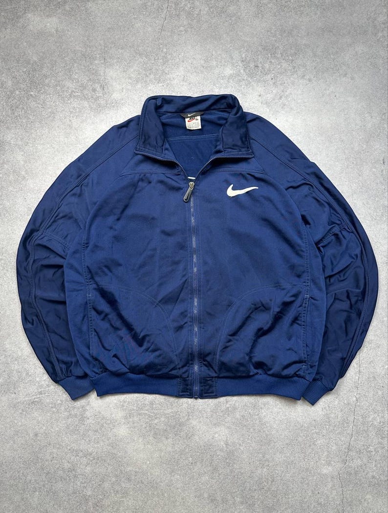 Pode incluir: Casaco de treino azul marinho com fecho de correr e log&oacute;tipo Nike branco no peito. Gola alta, punhos e bainha canelados. Uma pequena etiqueta Nike &eacute; vis&iacute;vel na gola.