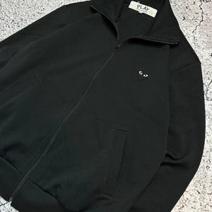 May include: Black zip-up jacket with a high collar and two front pockets. The jacket features a small heart-shaped logo with eyes on the chest. The label reads "PLAY".