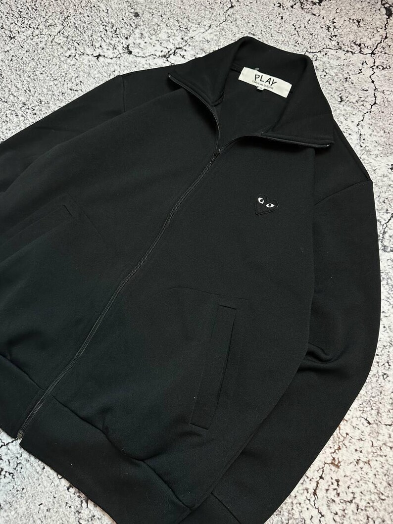May include: Black zip-up jacket with a high collar and two front pockets. The jacket features a small heart-shaped logo with eyes on the chest. The label reads "PLAY".