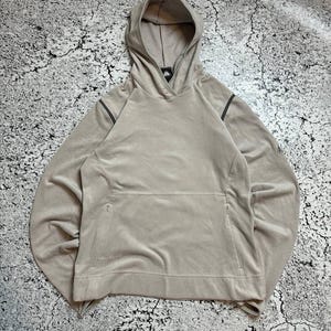 Puede incluir: Una sudadera con capucha de color beige claro con capucha con cordón y bolsillo tipo canguro. La sudadera tiene una textura suave y aterciopelada y presenta un pequeño logotipo en el pecho y rayas grises oscuras en las mangas.