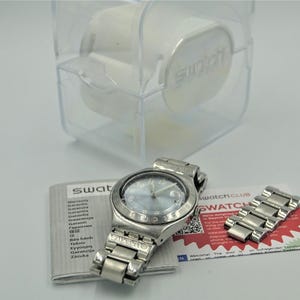 May include: A silver-toned Swatch watch with a light blue face and matching metal band, displayed with an extra link and paperwork. The watch is in its original clear plastic case, with the Swatch logo visible.