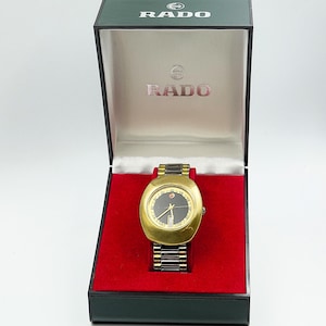 May include: A vintage Rado watch in its original box. The watch features a gold-toned case and a black dial with gold hands. The watch band is a combination of gold and black links. The watch is presented in a green and black box with the Rado logo.