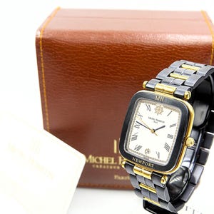 May include: A black and gold wristwatch with a square face and a black and gold band. The watch face is white with black Roman numerals and the text "MICHEL HERBELIN PARIS NEWPORT". A brown leather box is in the background.