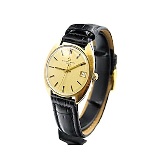 May include: A gold-tone wristwatch with a black leather band. The watch face is round and has a gold-tone bezel. The watch face is white with black markings and the text "ETERNA-MATIC 2000" printed on it.