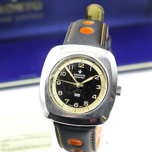 May include: A vintage Pronto Sportal GS wristwatch with a black dial and gold-colored numerals. The watch has a silver-colored square case and a black leather strap with orange accents. The watch is displayed in a blue box.