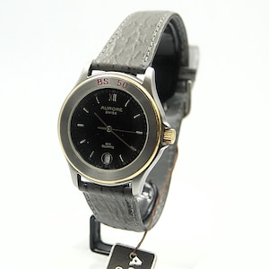 Puede incluir: Un reloj de pulsera plateado con esfera negra y bisel dorado. El reloj tiene una correa de cuero gris y el texto "AURORE SWISS" en la esfera. El reloj tiene una pantalla de fecha y está marcado "BS 50".