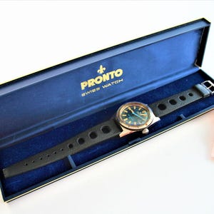 Op de afbeelding: Een vintage Pronto Swiss Watch in de originele presentatiedoos. Het horloge heeft een zwarte leren band met cirkelvormige uitsparingen en een zilverkleurige kast. De wijzerplaat heeft een turquoise kleur. De doos is marineblauw met gouden randen.