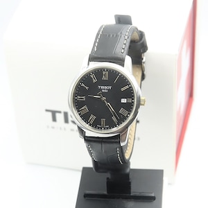 May include: A Tissot 1853 wristwatch featuring a round silver-tone case, a black dial with Roman numeral hour markers, and a date window at the three o'clock position. The watch includes a black leather strap with white stitching and a gold-tone crown.
