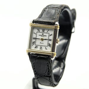 May include: A rectangular Michel Herbelin quartz watch with a white face, gold-tone bezel, and black leather band. The watch face features Roman numerals and the words "Paris" and "France". The watch is displayed against a white background.