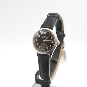 Puede incluir: Un reloj de pulsera Oris vintage con esfera negra y caja plateada. El reloj tiene una correa de cuero negro y una pequeña subesfera de segundos. La esfera muestra números arábigos y el texto "ORIS WATERPROOF ANTI-SHOCK".