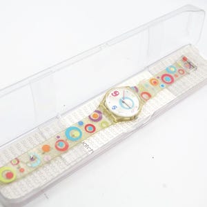 May include: A Swatch watch with a transparent band and a white face. The band is decorated with colorful circles. The watch is in a clear plastic case. The watch face has the numbers 3, 6, and 9 in red.