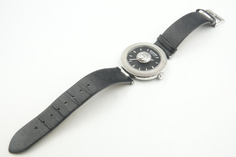 Puede incluir: Un reloj de pulsera con correa negra texturizada y caja redonda plateada. La esfera negra presenta un emblema central de cabeza de le&oacute;n plateado, manecillas plateadas y el texto Versus Versace. El bisel tiene una textura finamente moleteada.