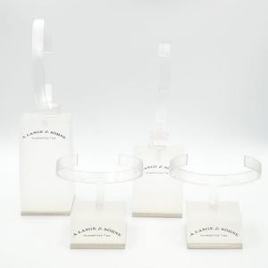 May include: Three clear plastic watch display stands with a white base. Each stand has a curved arm to hold a watch and a label that reads "A. Lange & Söhne Glashütte i/Sa."