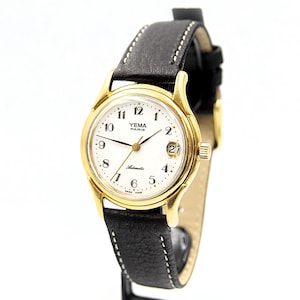 May include: A gold-toned Yema Paris automatic watch with a white face, black hands, and a date display. The watch has a black leather band with white stitching. The watch face displays the numbers 1 through 12.