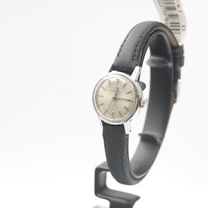 May include: A silver wristwatch with a white face and black leather strap. The watch is on a black stand.