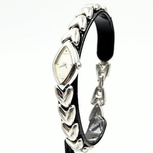 Puede incluir: Un reloj Seiko plateado con esfera blanca y correa de cuero negro. La correa del reloj presenta eslabones en forma de corazón y un cierre plateado. El reloj tiene una esfera en forma de diamante y agujas plateadas.