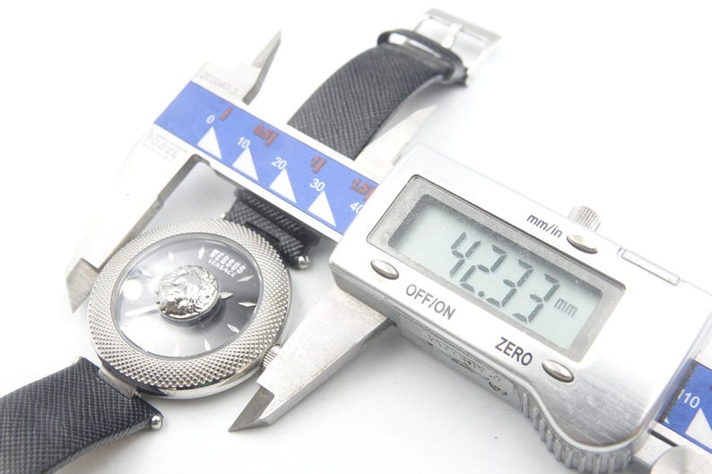 Puede incluir: Un reloj de pulsera Versus Versace con esfera negra, emblema de cabeza de le&oacute;n plateado y bisel plateado texturizado. Un calibre digital mide la caja del reloj en 42,33 mm. El reloj cuenta con una correa negra texturizada e &iacute;ndices plateados.