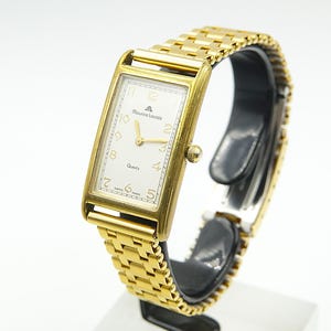 Peut inclure: Une montre-bracelet rectangulaire dorée avec un cadran blanc. La montre est dotée d'un bracelet doré et d'un petit cadran. Le cadran affiche la marque "Maurice Lacroix" et le mot "Quartz". La montre est présentée sur un support noir.